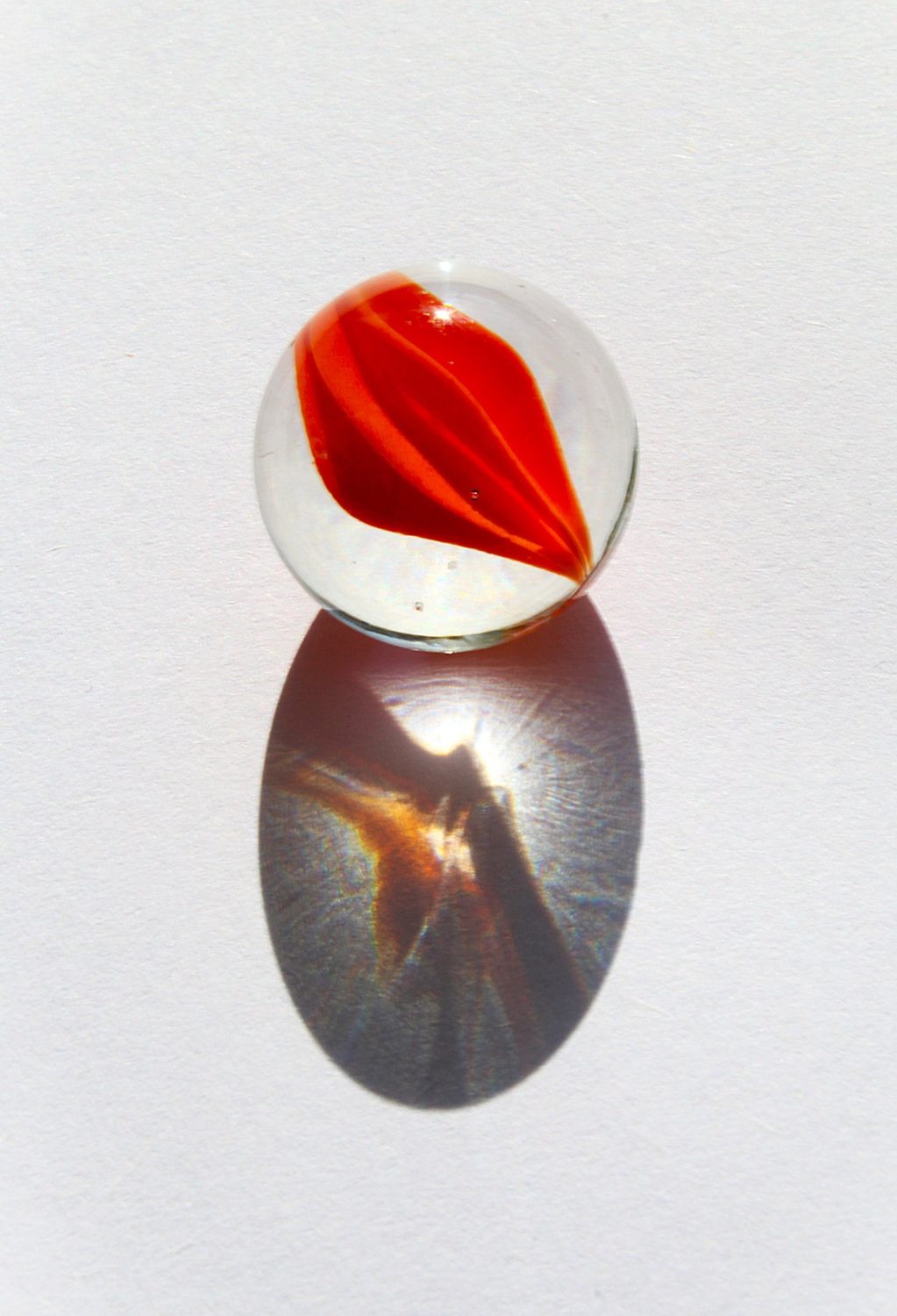 Clear glass marble with a red swirl, casting caustics onto white paper